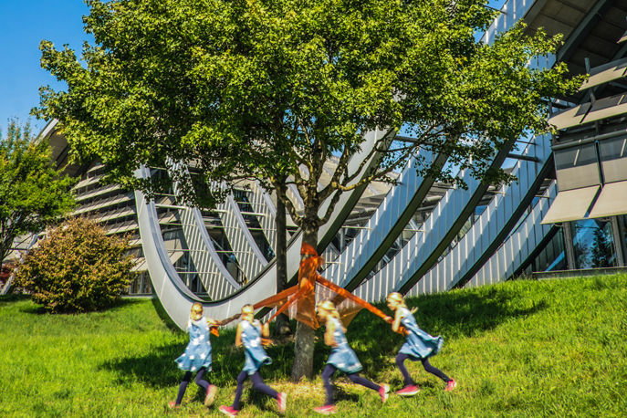 Ein Mädchen, dass mit einem Tuch rund um einen grünen Baum vor dem Zentrum Paul Klee rennt