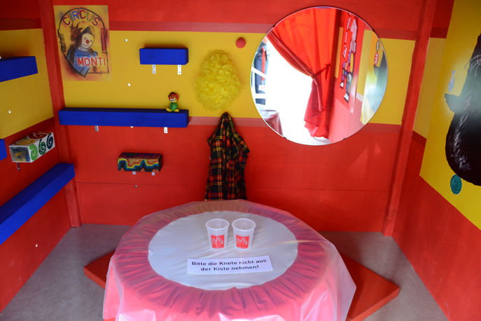 Blick in eine Kiste rot/gelbe Kiste in welcher ein runder Tisch steht und ein Spiegel an der Wand hängt