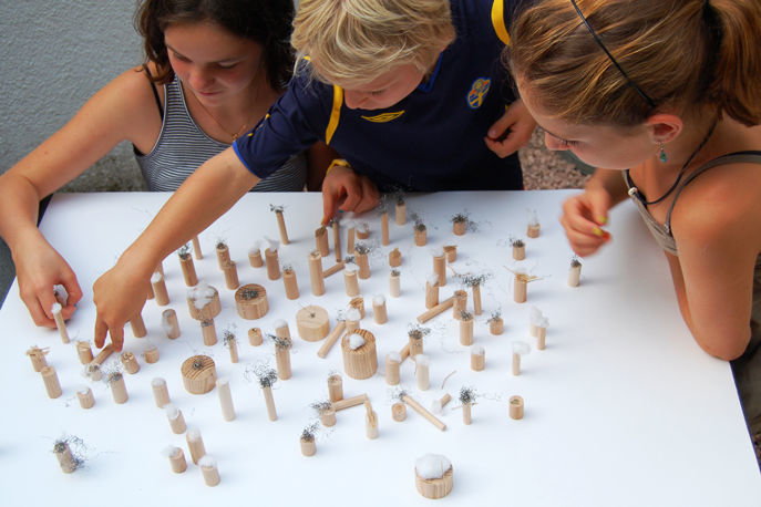 Drei Kinder betrachten  kleine Engel aus Holz welche auf einem weissen Brett platziert sind