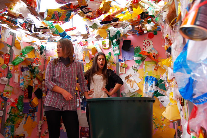 Zwei Mädchen welche in einem Raum voll Müll stehen in welchem sich eine leere Mülltonne befindet